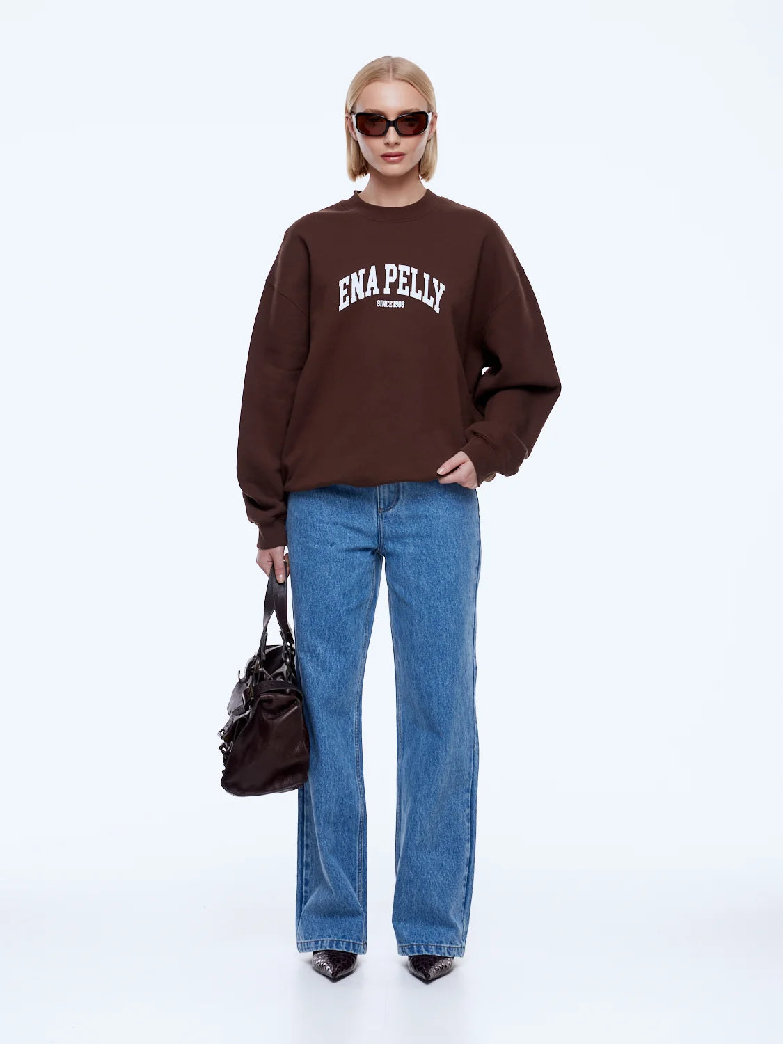 A model stands against a light gray studio background wearing a dark brown sweatshirt with 'ENA PELLY' printed in white across the chest, paired with blue jeans, black pointed shoes, sunglasses, and carrying a dark brown handbag.
