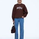 A model stands against a light gray studio background wearing a dark brown sweatshirt with 'ENA PELLY' printed in white across the chest, paired with blue jeans, black pointed shoes, sunglasses, and carrying a dark brown handbag.