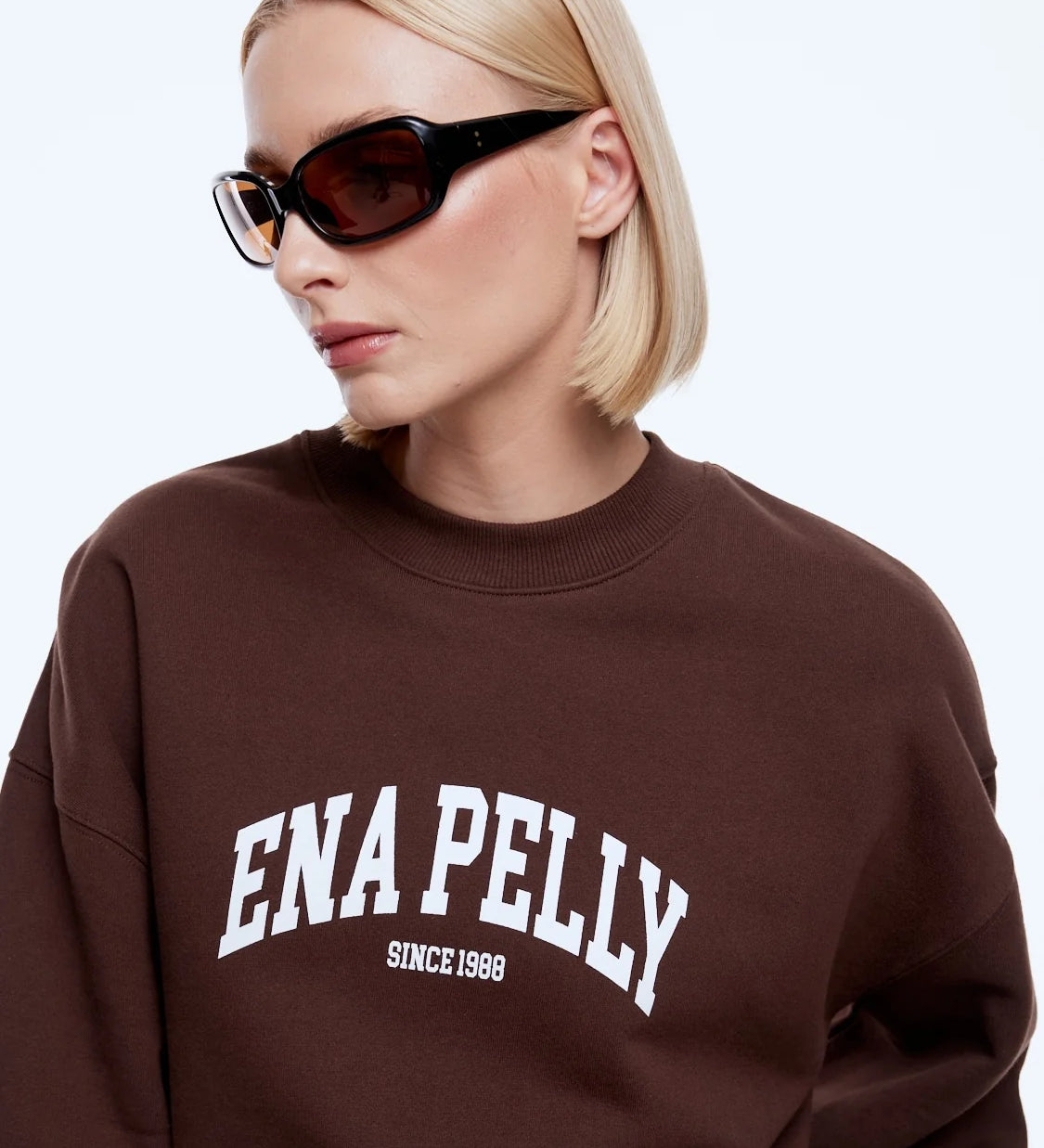 A blonde woman wearing a brown crewneck sweatshirt with 'ENA PELLY SINCE 1988' printed in white on the front, paired with black sunglasses, against a light gray background.