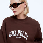 A blonde woman wearing a brown crewneck sweatshirt with 'ENA PELLY SINCE 1988' printed in white on the front, paired with black sunglasses, against a light gray background.