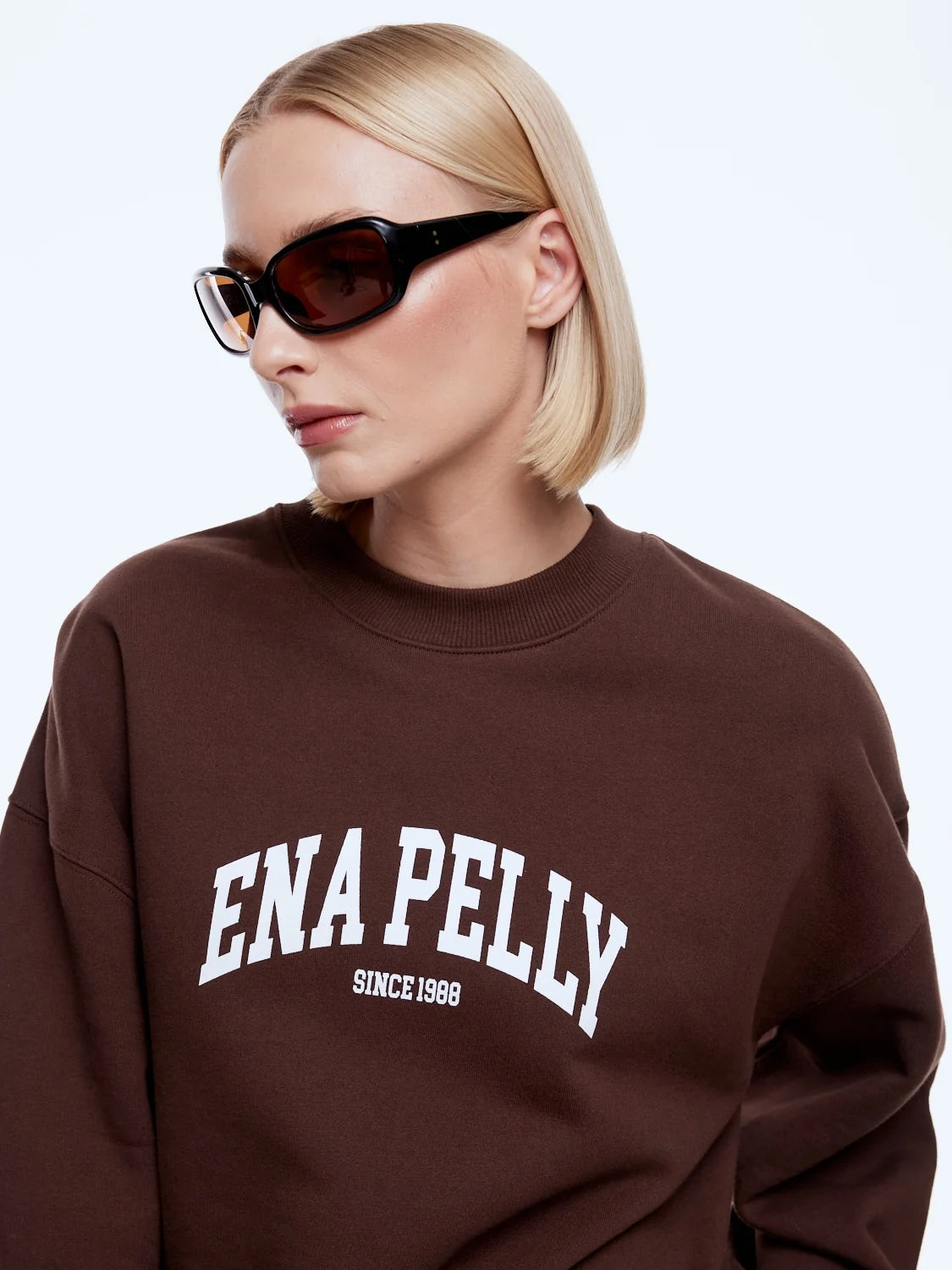 A blonde woman wearing a brown crewneck sweatshirt with 'ENA PELLY SINCE 1988' printed in white on the front, paired with black sunglasses, against a light gray background.