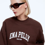 A blonde woman wearing a brown crewneck sweatshirt with 'ENA PELLY SINCE 1988' printed in white on the front, paired with black sunglasses, against a light gray background.