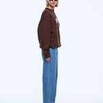 A model wearing a brown crewneck sweatshirt with a white graphic on the chest, paired with wide-leg blue jeans and black pointed shoes, photographed against a solid white studio background.