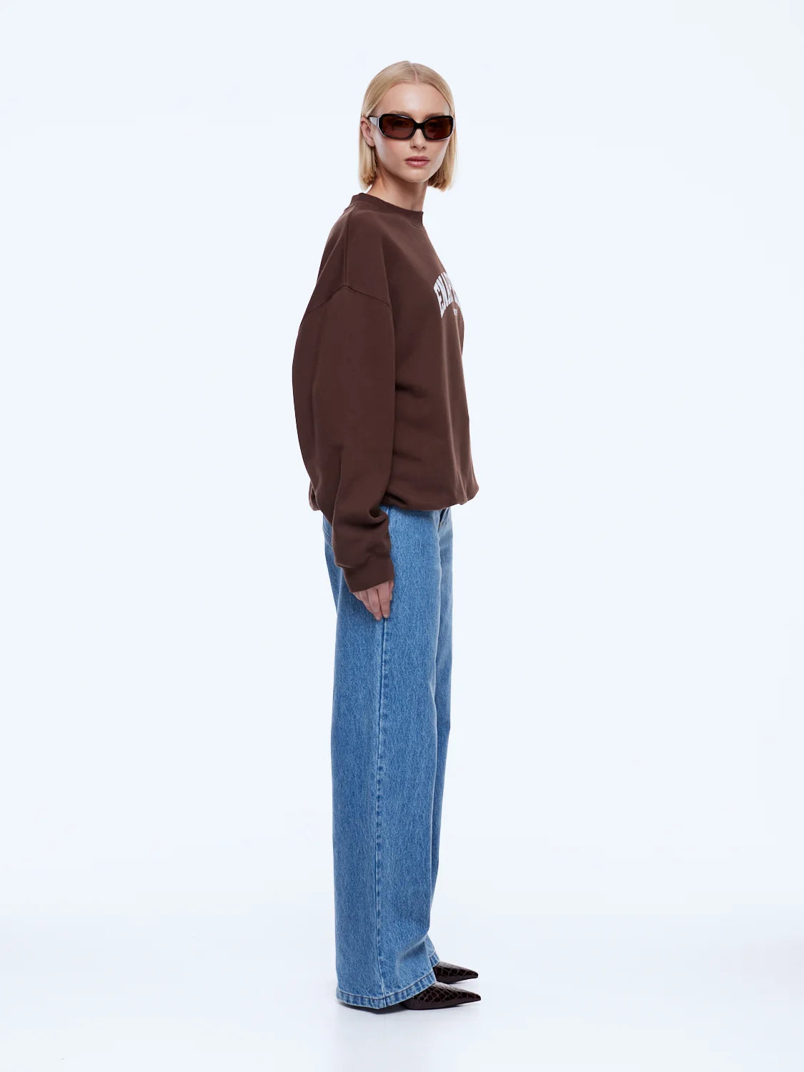 A model wearing a brown crewneck sweatshirt with a white graphic on the chest, paired with wide-leg blue jeans and black pointed shoes, photographed against a solid white studio background.
