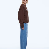 A model wearing a brown crewneck sweatshirt with a white graphic on the chest, paired with wide-leg blue jeans and black pointed shoes, photographed against a solid white studio background.