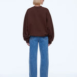 A model viewed from the back wearing a dark brown crewneck sweatshirt and wide-leg blue jeans against a plain white studio background.