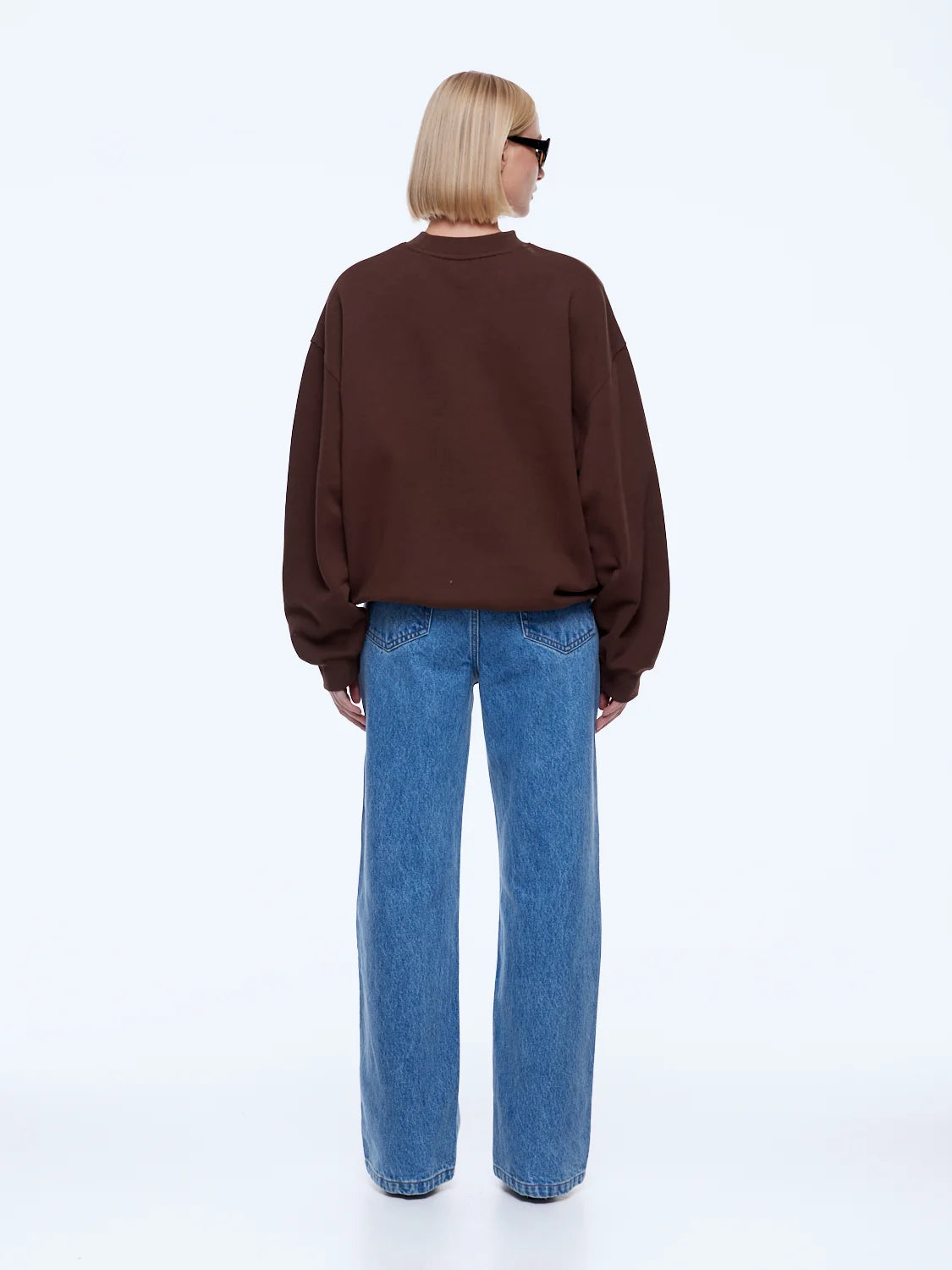 A model viewed from the back wearing a dark brown crewneck sweatshirt and wide-leg blue jeans against a plain white studio background.