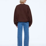 A model viewed from the back wearing a dark brown crewneck sweatshirt and wide-leg blue jeans against a plain white studio background.