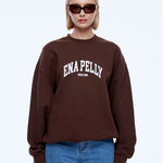 A blonde model wearing a dark brown crewneck sweatshirt with 'ENA PELLY' printed in white across the chest, paired with blue jeans and dark sunglasses, against a light gray background.