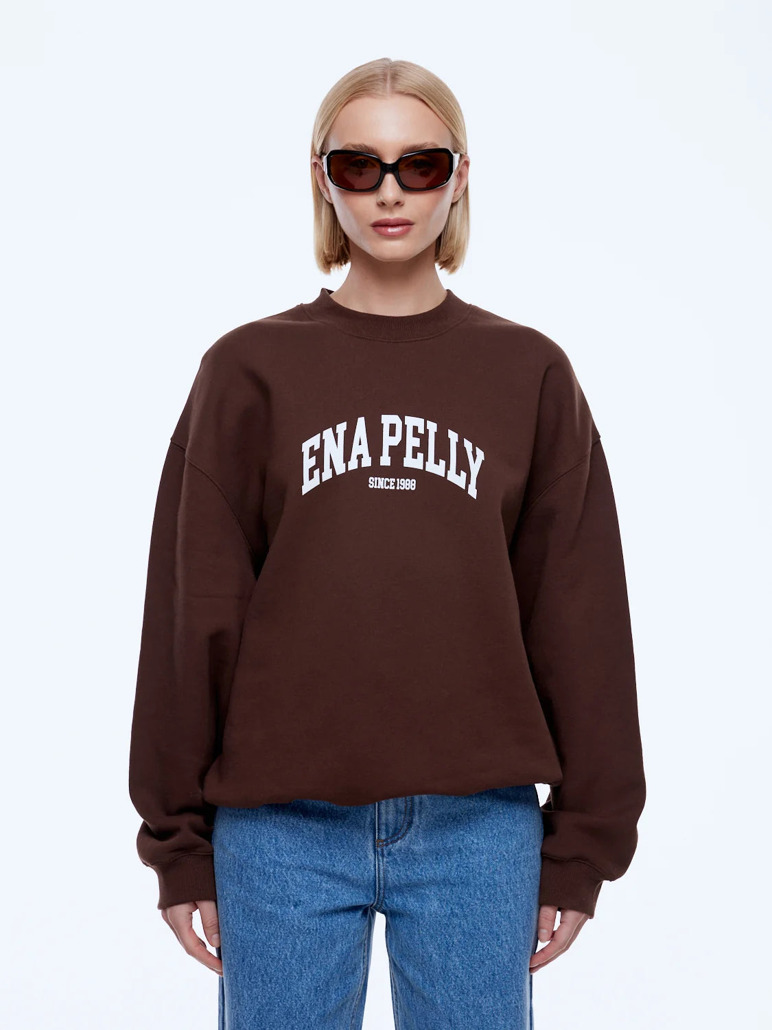 A blonde model wearing a dark brown crewneck sweatshirt with 'ENA PELLY' printed in white across the chest, paired with blue jeans and dark sunglasses, against a light gray background.