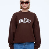 A blonde model wearing a dark brown crewneck sweatshirt with 'ENA PELLY' printed in white across the chest, paired with blue jeans and dark sunglasses, against a light gray background.