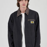A male model wearing a black Rollas work jacket with a brown corduroy collar, layered over a white t-shirt and brown pants, against a light gray background.