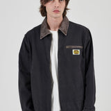 A male model wearing a black Rollas work jacket with a brown corduroy collar, layered over a white t-shirt and brown pants, against a light gray background.