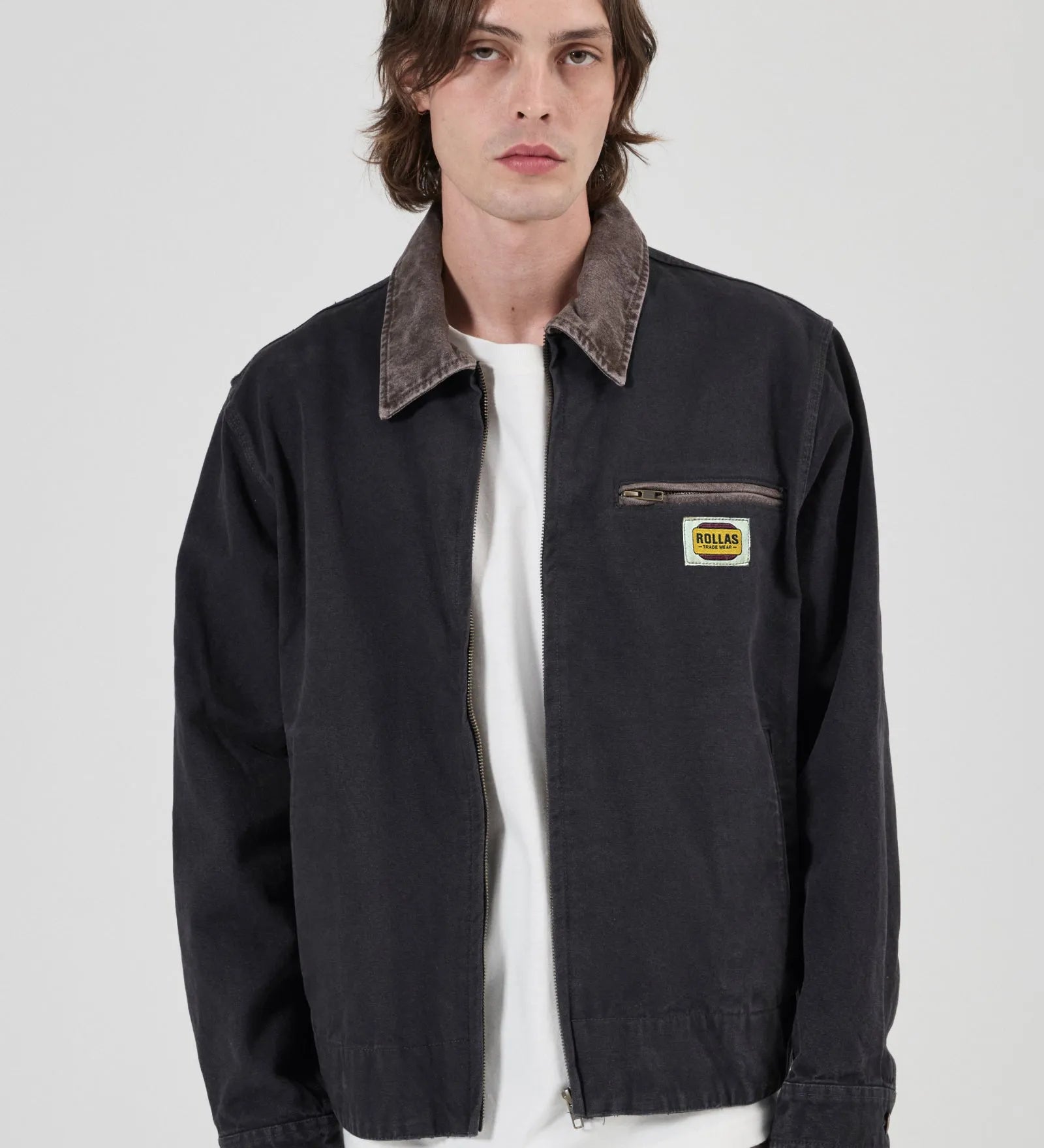 A male model wearing a black Rollas work jacket with a brown corduroy collar, layered over a white t-shirt and brown pants, against a light gray background.