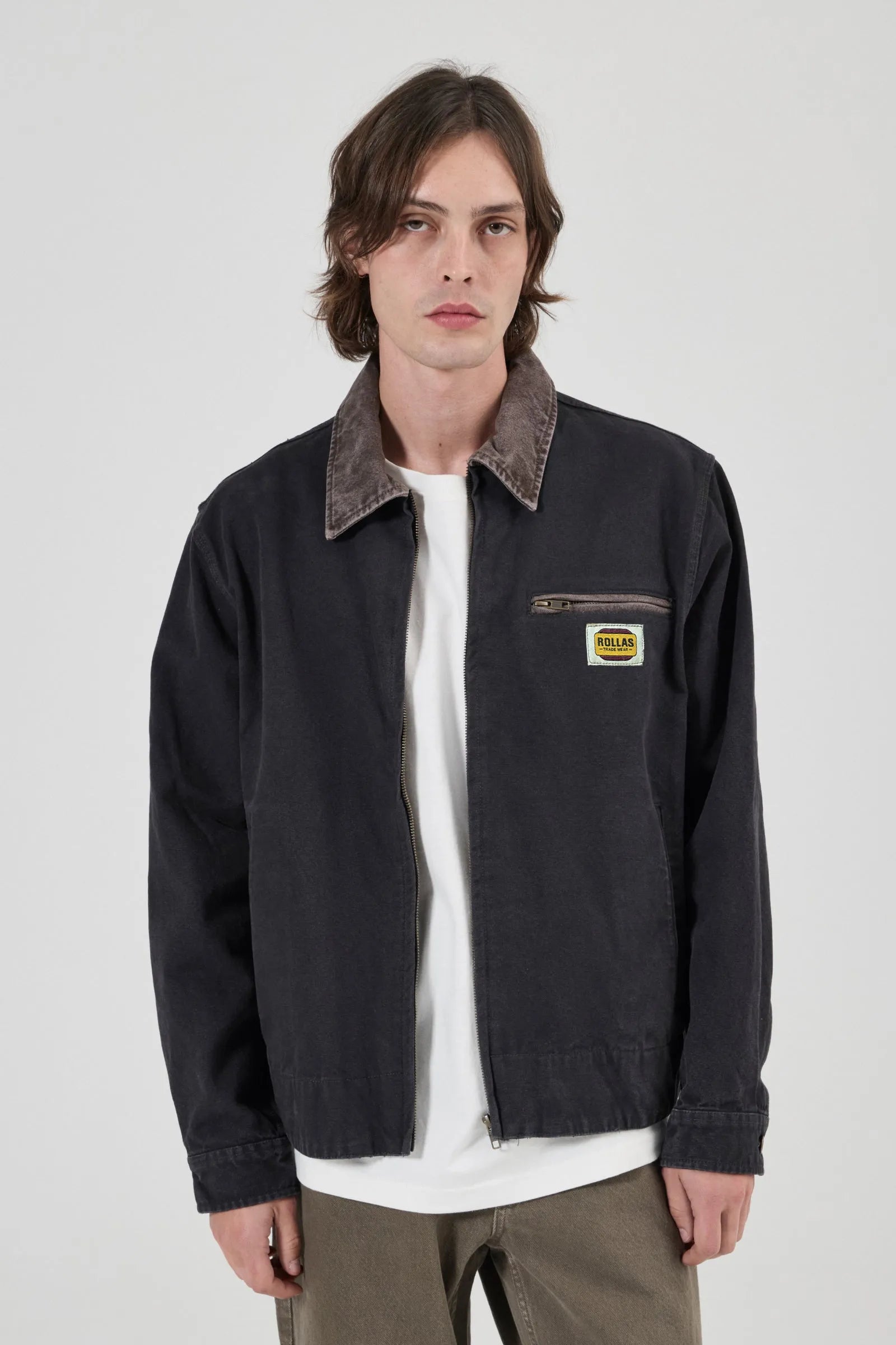 A male model wearing a black Rollas work jacket with a brown corduroy collar, layered over a white t-shirt and brown pants, against a light gray background.