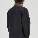 A person shown from the back wearing a dark charcoal denim jacket over a collared shirt and olive green pants, photographed against a plain light gray studio background.