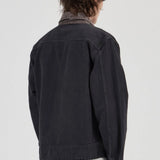 A person shown from the back wearing a dark charcoal denim jacket over a collared shirt and olive green pants, photographed against a plain light gray studio background.