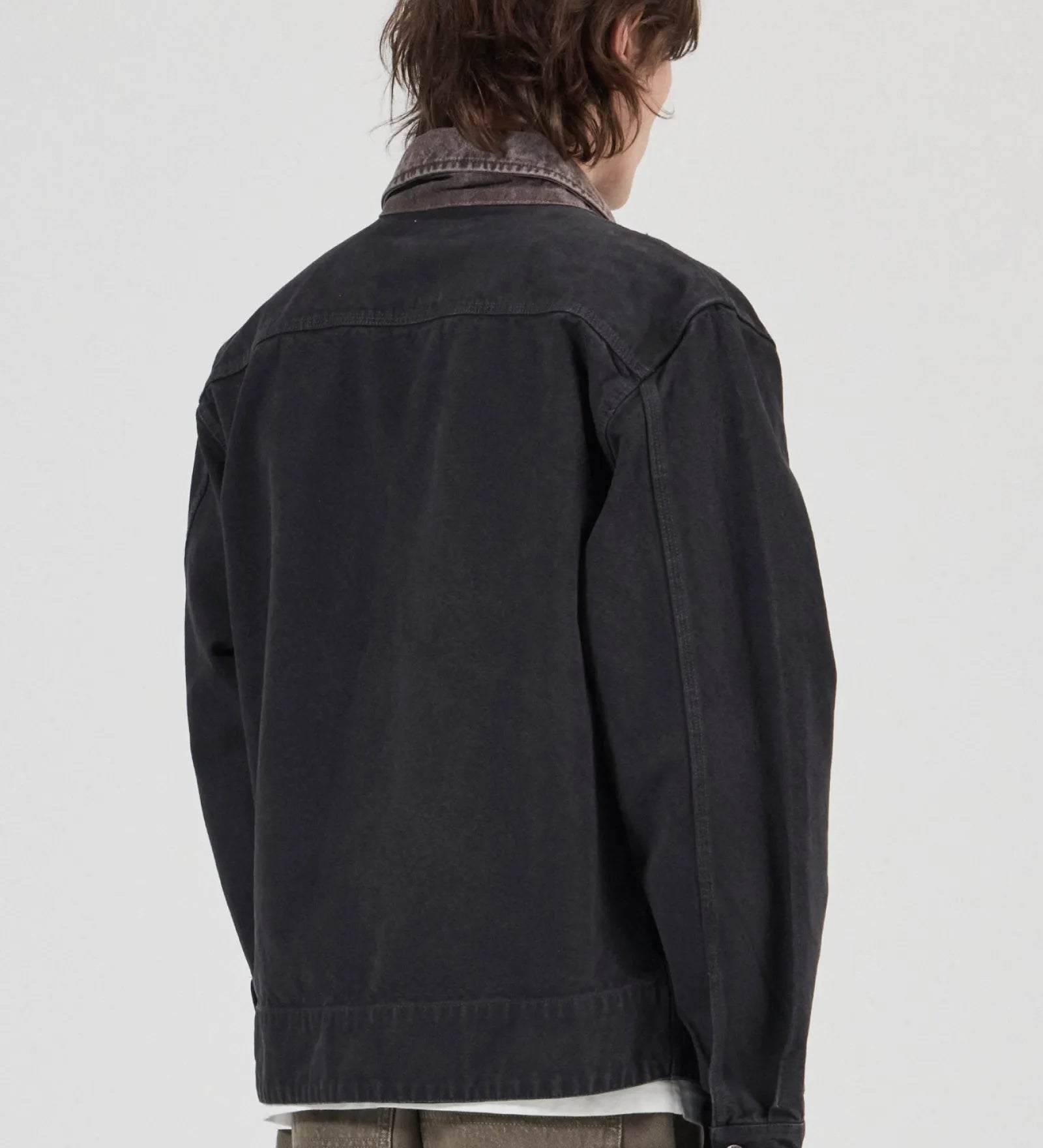 A person shown from the back wearing a dark charcoal denim jacket over a collared shirt and olive green pants, photographed against a plain light gray studio background.
