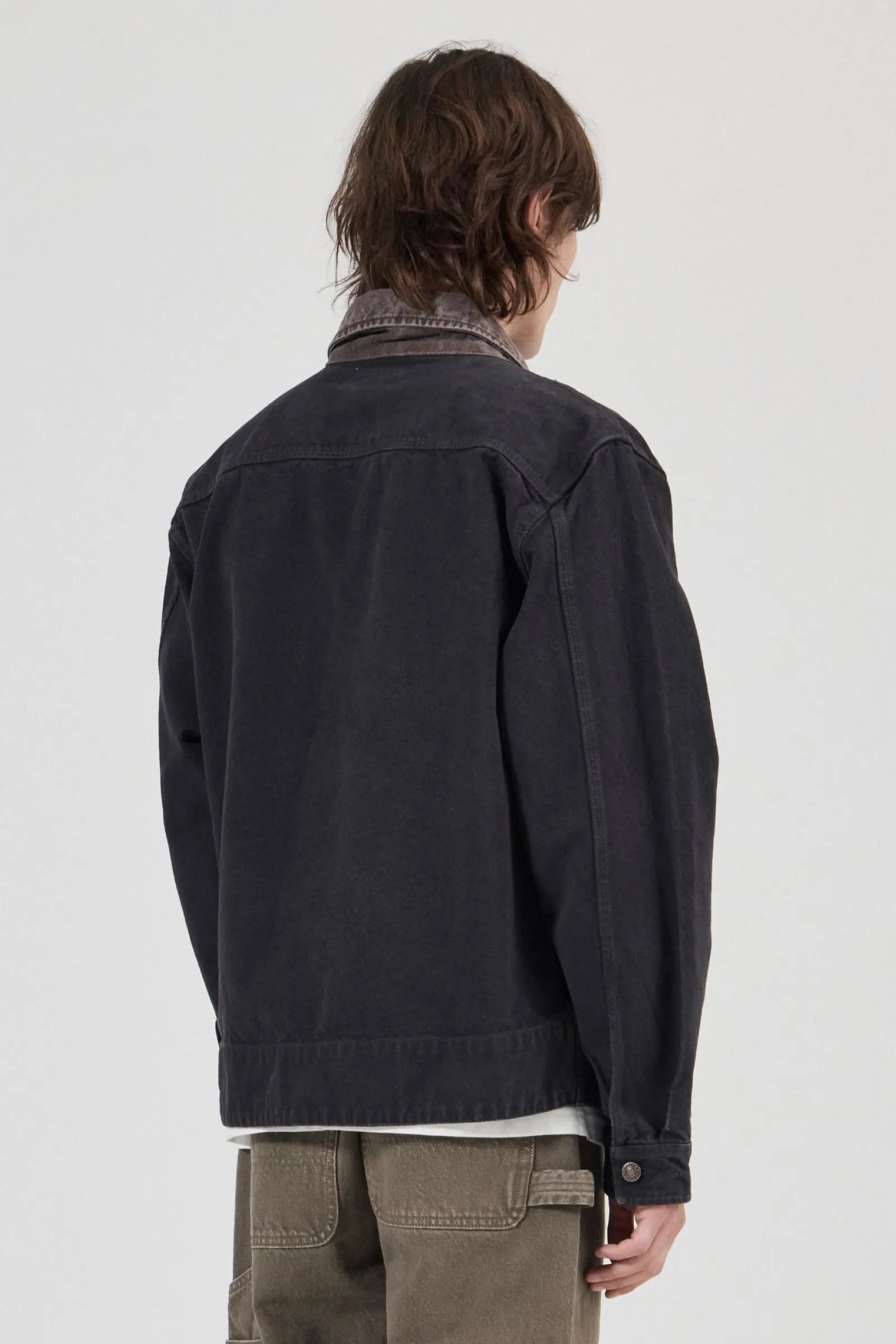 A person shown from the back wearing a dark charcoal denim jacket over a collared shirt and olive green pants, photographed against a plain light gray studio background.