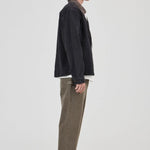 A model shown in profile wearing a dark charcoal jacket, olive green corduroy trousers, a white shirt, and Adidas sneakers against a plain light gray studio background.