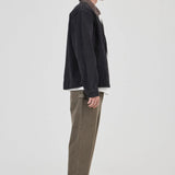 A model shown in profile wearing a dark charcoal jacket, olive green corduroy trousers, a white shirt, and Adidas sneakers against a plain light gray studio background.