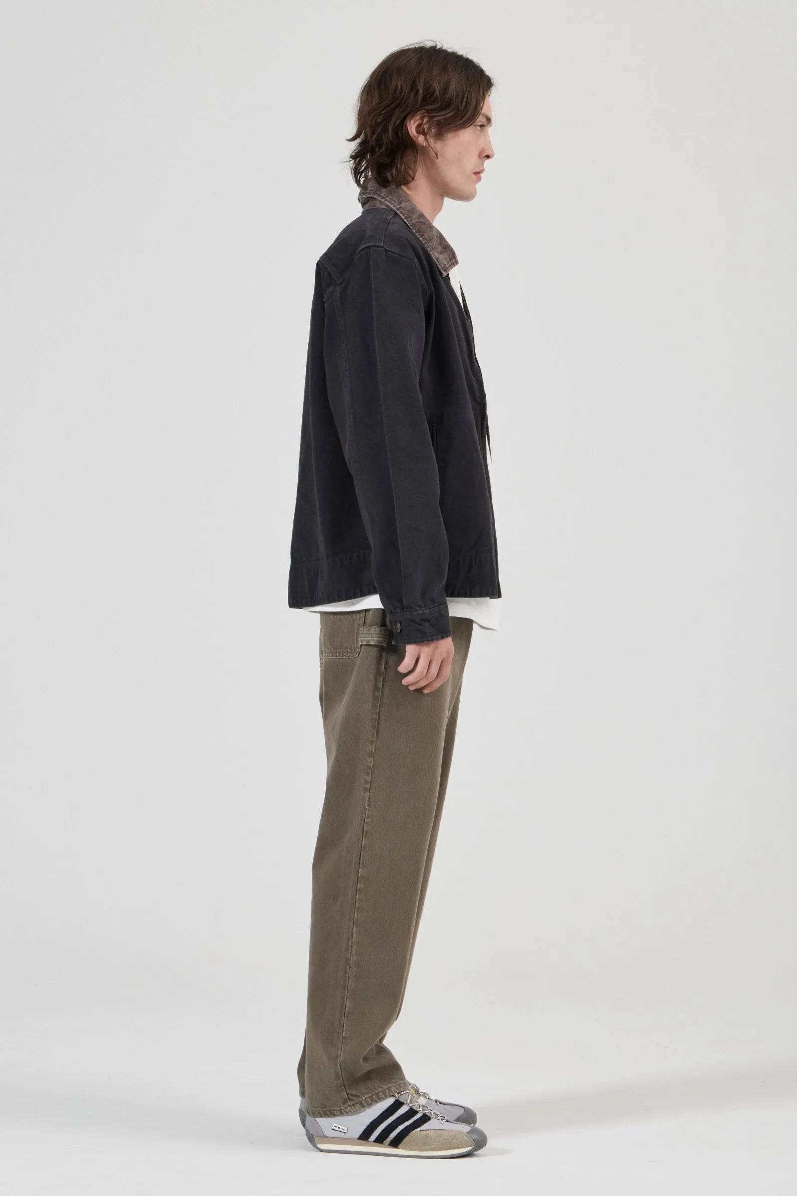 A model shown in profile wearing a dark charcoal jacket, olive green corduroy trousers, a white shirt, and Adidas sneakers against a plain light gray studio background.