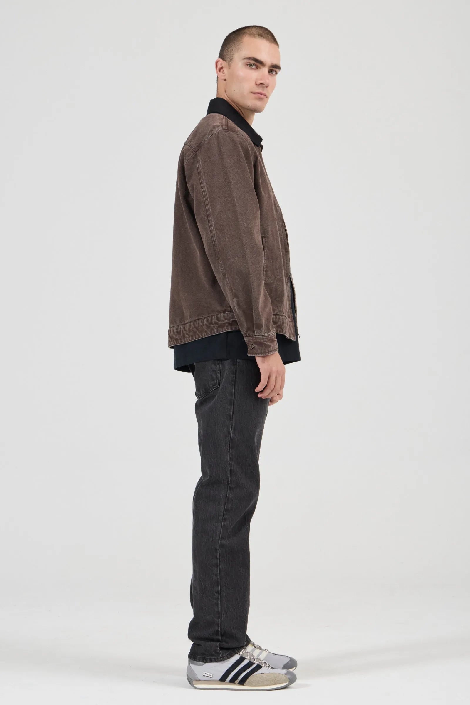 Man wearing a brown jacket and dark jeans standing against a white background