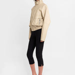 A model is shown from the side wearing a beige faux-leather jacket with a high collar, black cropped leggings, and clear high-heeled sandals with a black and white zebra print strap.