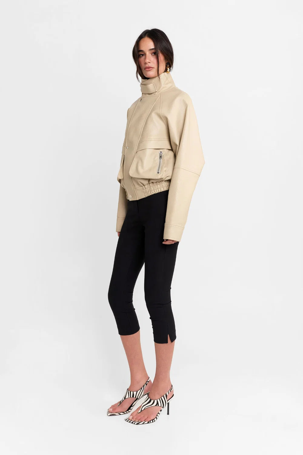 A model is shown from the side wearing a beige faux-leather jacket with a high collar, black cropped leggings, and clear high-heeled sandals with a black and white zebra print strap.