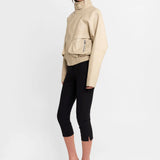 A model is shown from the side wearing a beige faux-leather jacket with a high collar, black cropped leggings, and clear high-heeled sandals with a black and white zebra print strap.