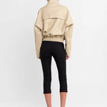 A model viewed from the back wearing a beige cropped jacket with a high collar, black cropped pants, and strappy high-heeled sandals, set against a plain white studio background.