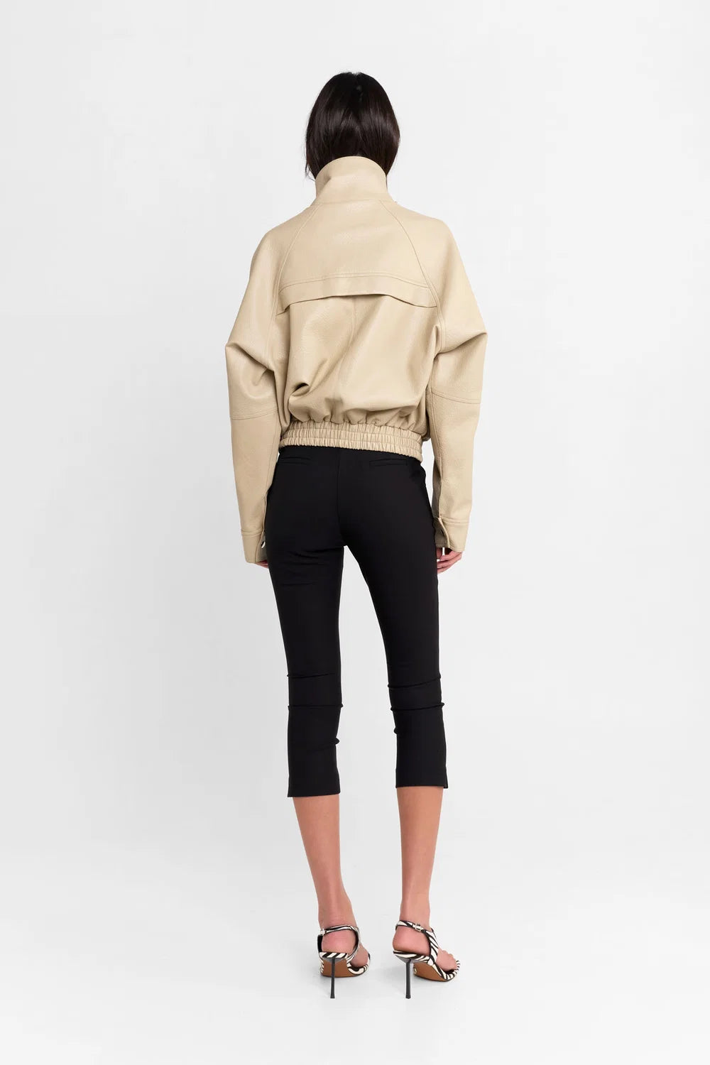 A model viewed from the back wearing a beige cropped jacket with a high collar, black cropped pants, and strappy high-heeled sandals, set against a plain white studio background.