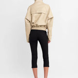 A model viewed from the back wearing a beige cropped jacket with a high collar, black cropped pants, and strappy high-heeled sandals, set against a plain white studio background.