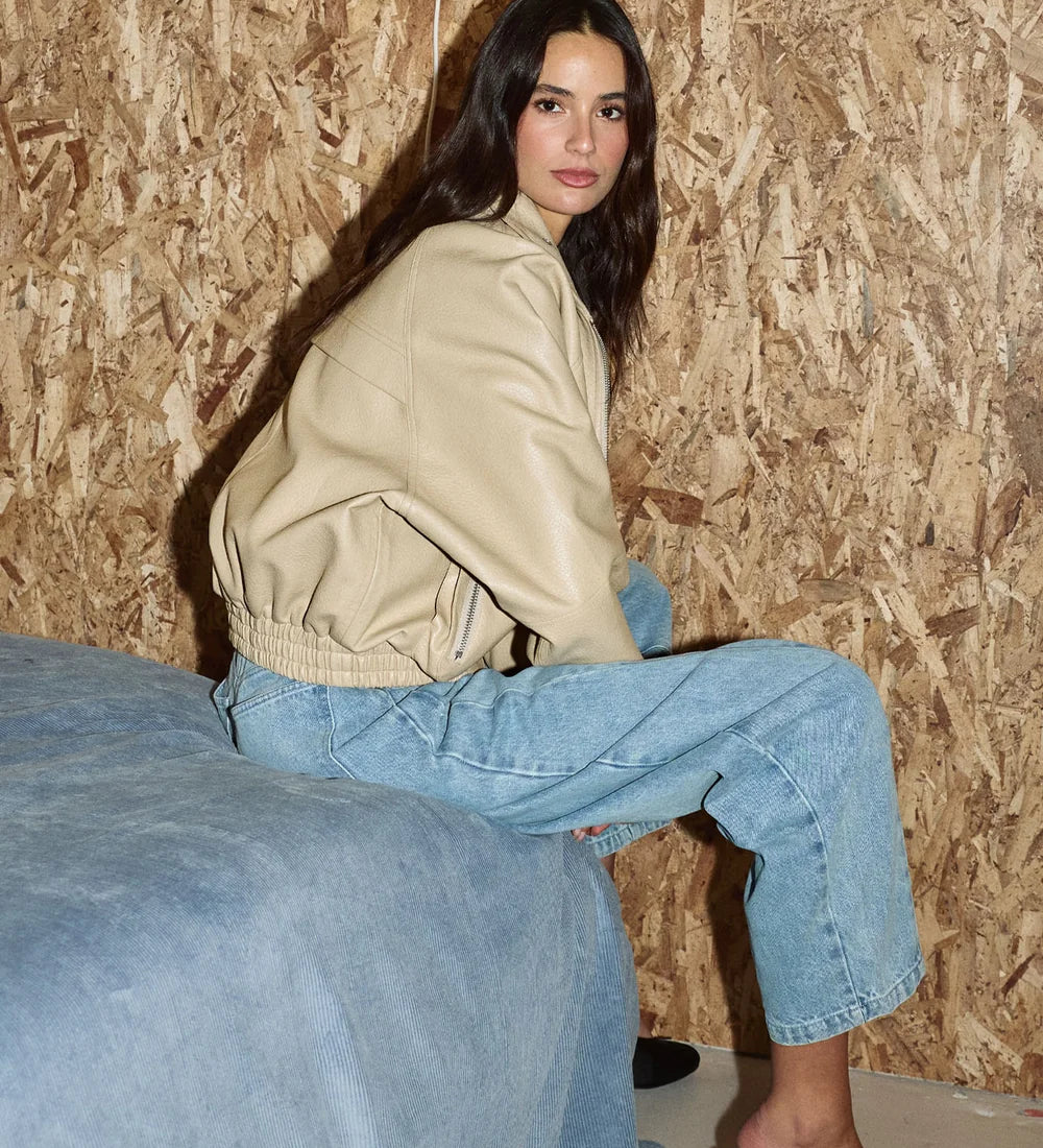 A woman with long dark hair sits on the edge of a blue bed, wearing a beige leather-look jacket, light-wash wide-leg jeans, and black high heels against an OSB wood-paneled wall.