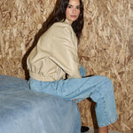 A woman with long dark hair sits on the edge of a blue bed, wearing a beige leather-look jacket, light-wash wide-leg jeans, and black high heels against an OSB wood-paneled wall.