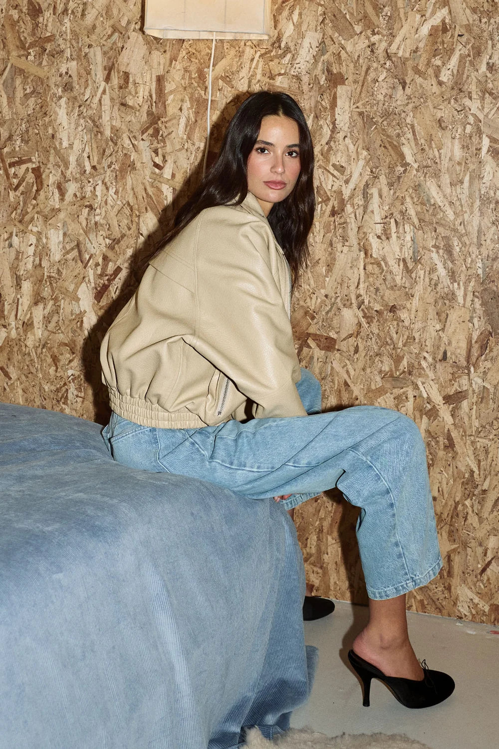A woman with long dark hair sits on the edge of a blue bed, wearing a beige leather-look jacket, light-wash wide-leg jeans, and black high heels against an OSB wood-paneled wall.