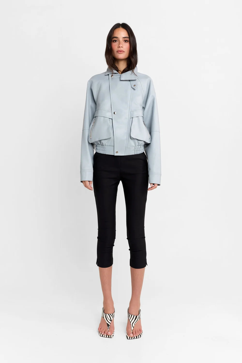 A model stands against a plain white studio background wearing a light blue cropped jacket, black cropped pants, and black-and-white striped high-heeled sandals.
