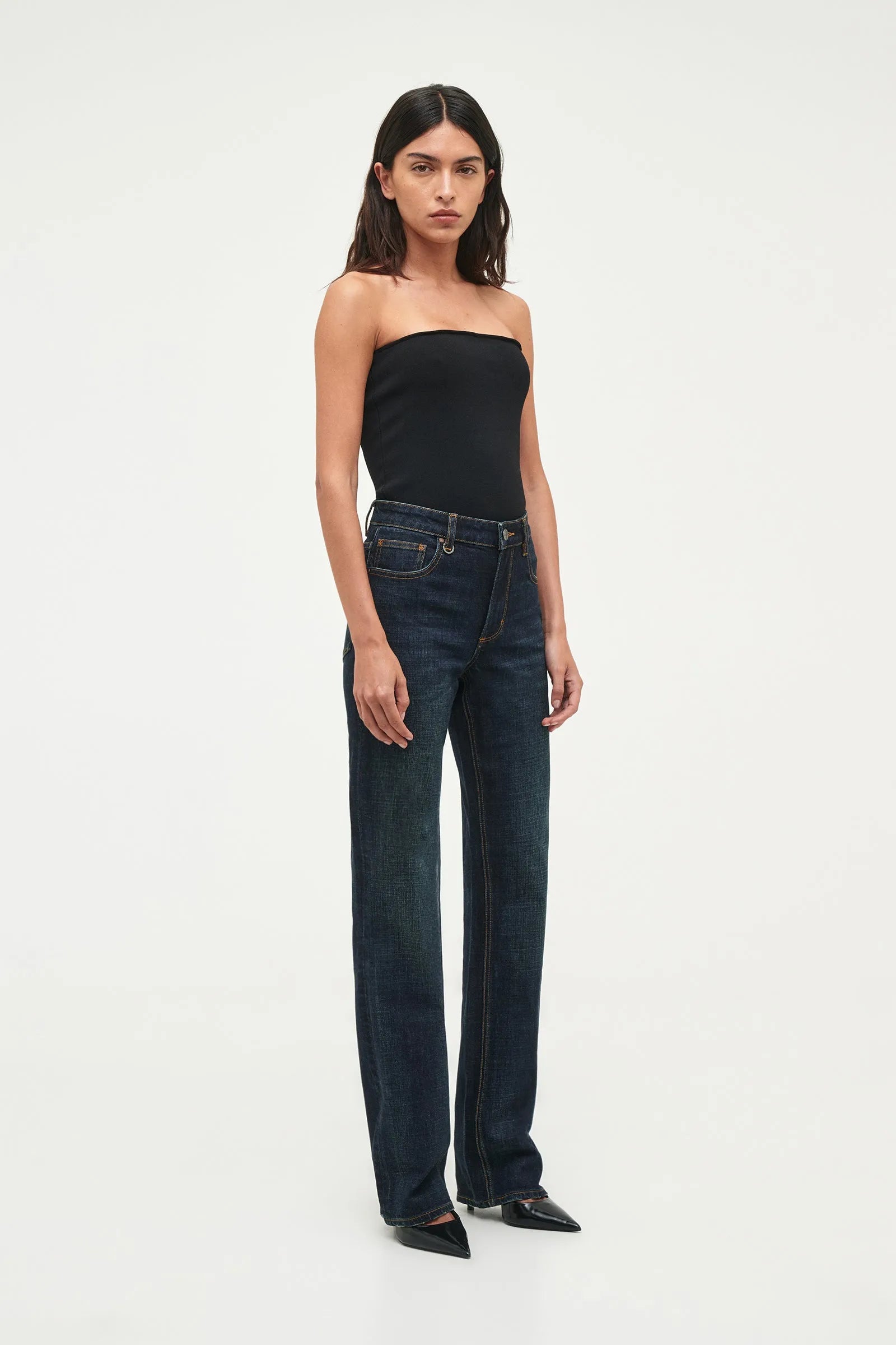 A model wearing dark wash, straight-leg jeans and a black strapless top, standing against a plain white studio background.