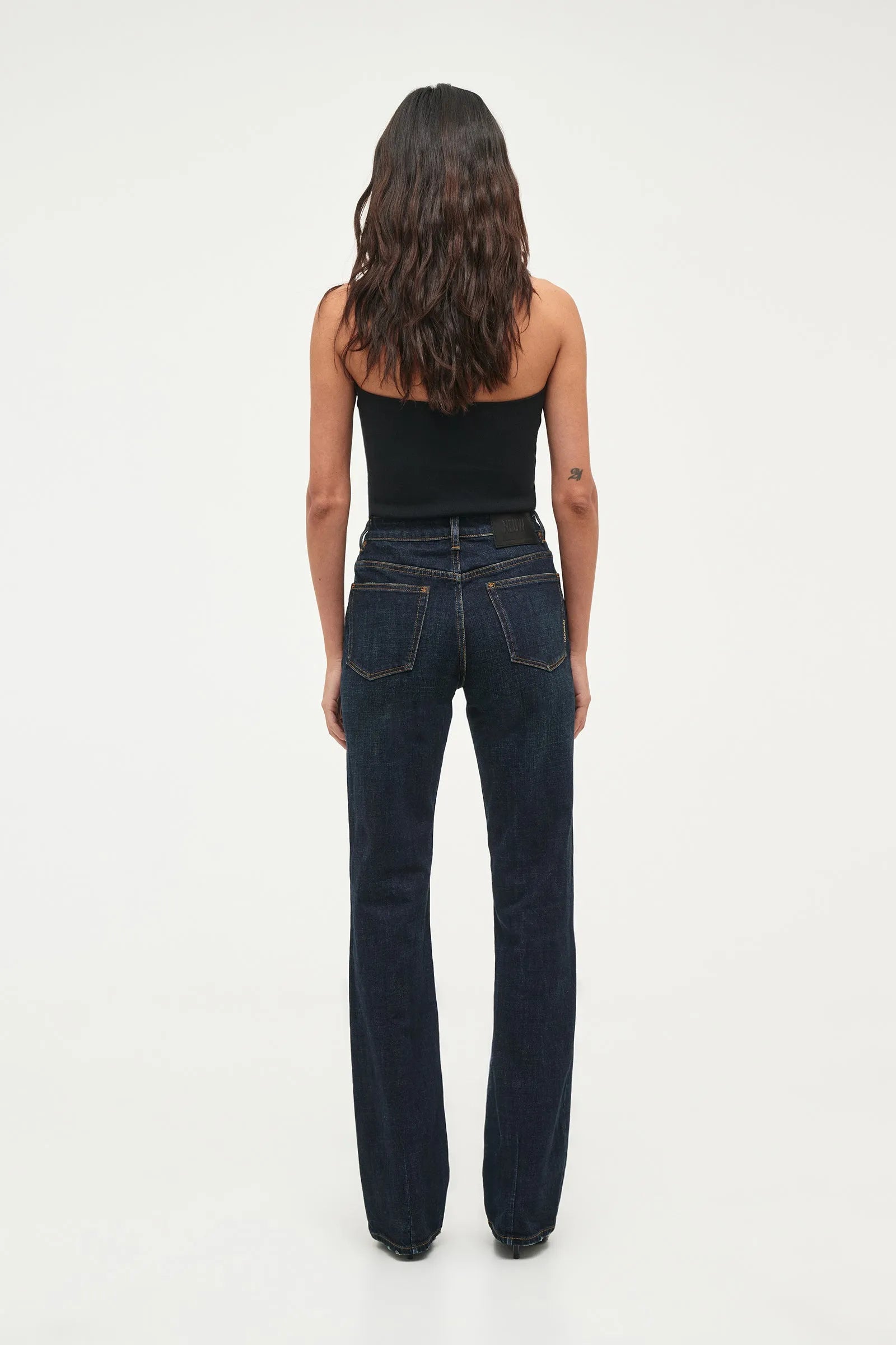 A woman viewed from behind wearing dark blue straight-leg jeans and a black strapless top against a plain white studio background.