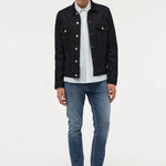 A male model stands against a light gray studio background wearing a dark denim jacket over a white striped button-up shirt, blue jeans, and black shoes.