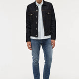 A male model stands against a light gray studio background wearing a dark denim jacket over a white striped button-up shirt, blue jeans, and black shoes.