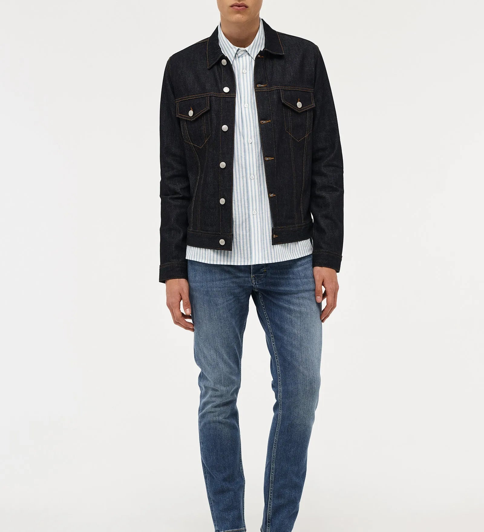 A male model stands against a light gray studio background wearing a dark denim jacket over a white striped button-up shirt, blue jeans, and black shoes.