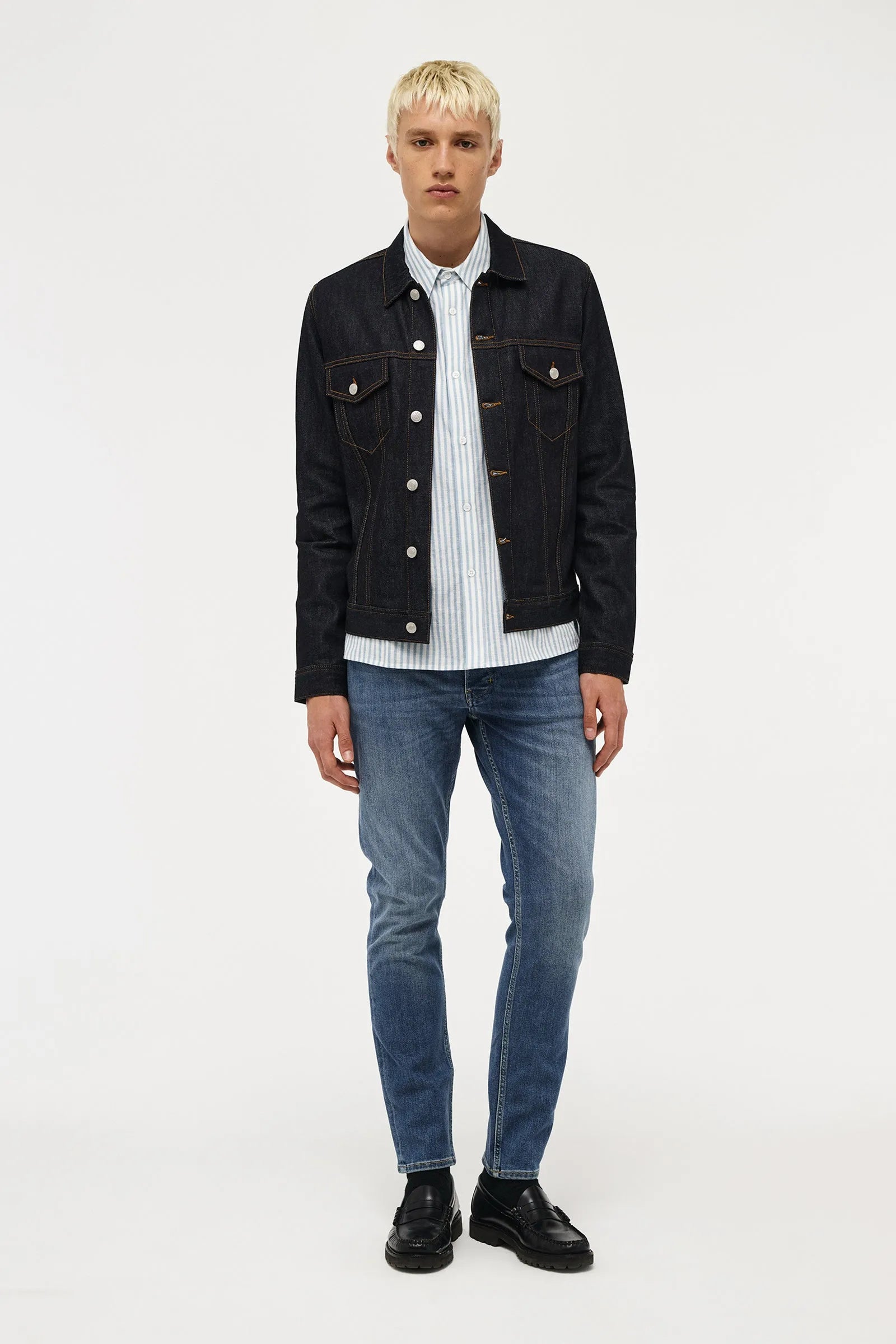 A male model stands against a light gray studio background wearing a dark denim jacket over a white striped button-up shirt, blue jeans, and black shoes.