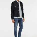 A male model with blonde hair stands against a light gray studio background wearing a black denim jacket over a white and blue striped button-down shirt, blue jeans, and black leather shoes.