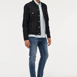 A male model with blonde hair stands against a light gray studio background wearing a black denim jacket over a white and blue striped button-down shirt, blue jeans, and black leather shoes.