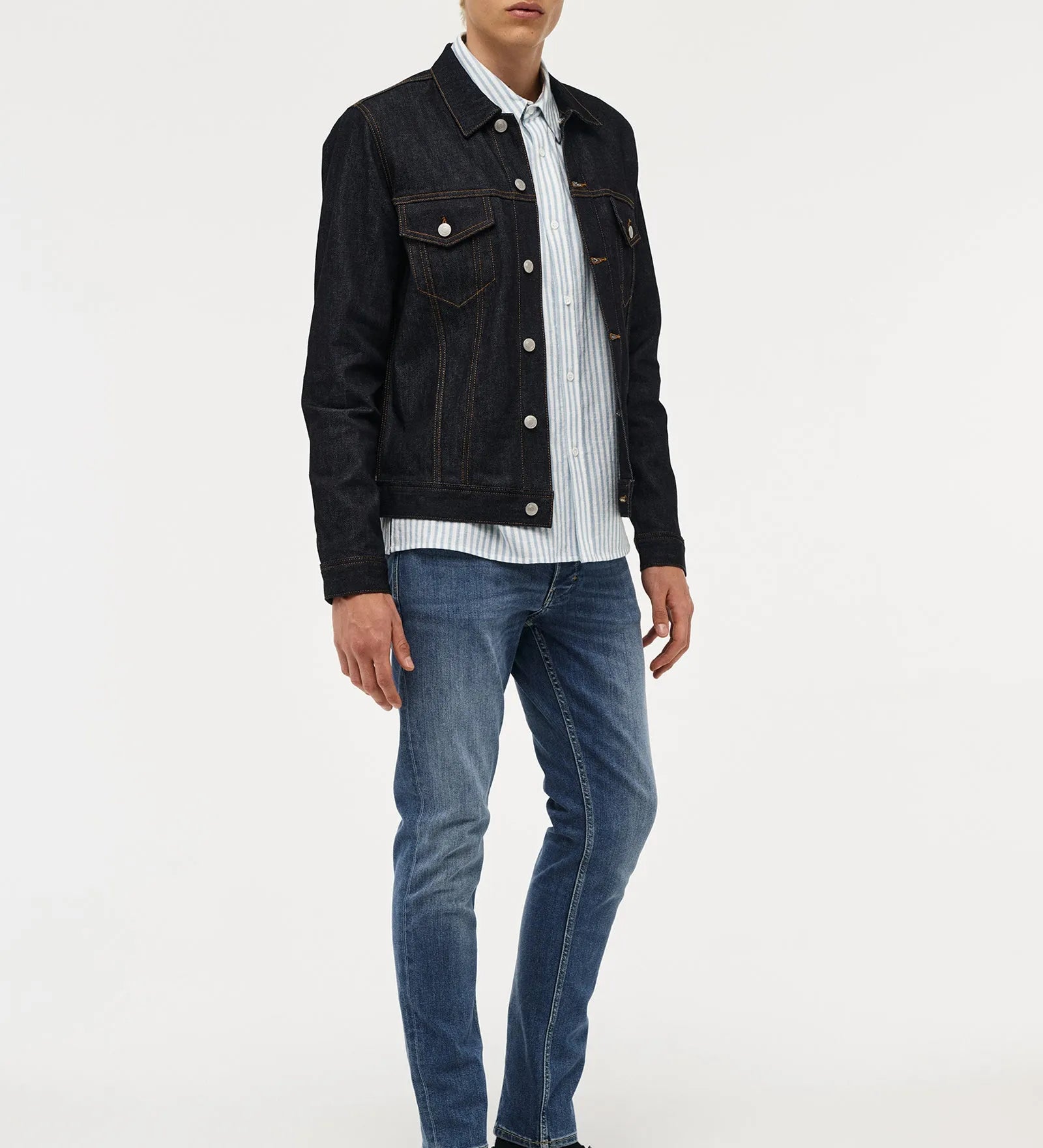 A male model with blonde hair stands against a light gray studio background wearing a black denim jacket over a white and blue striped button-down shirt, blue jeans, and black leather shoes.
