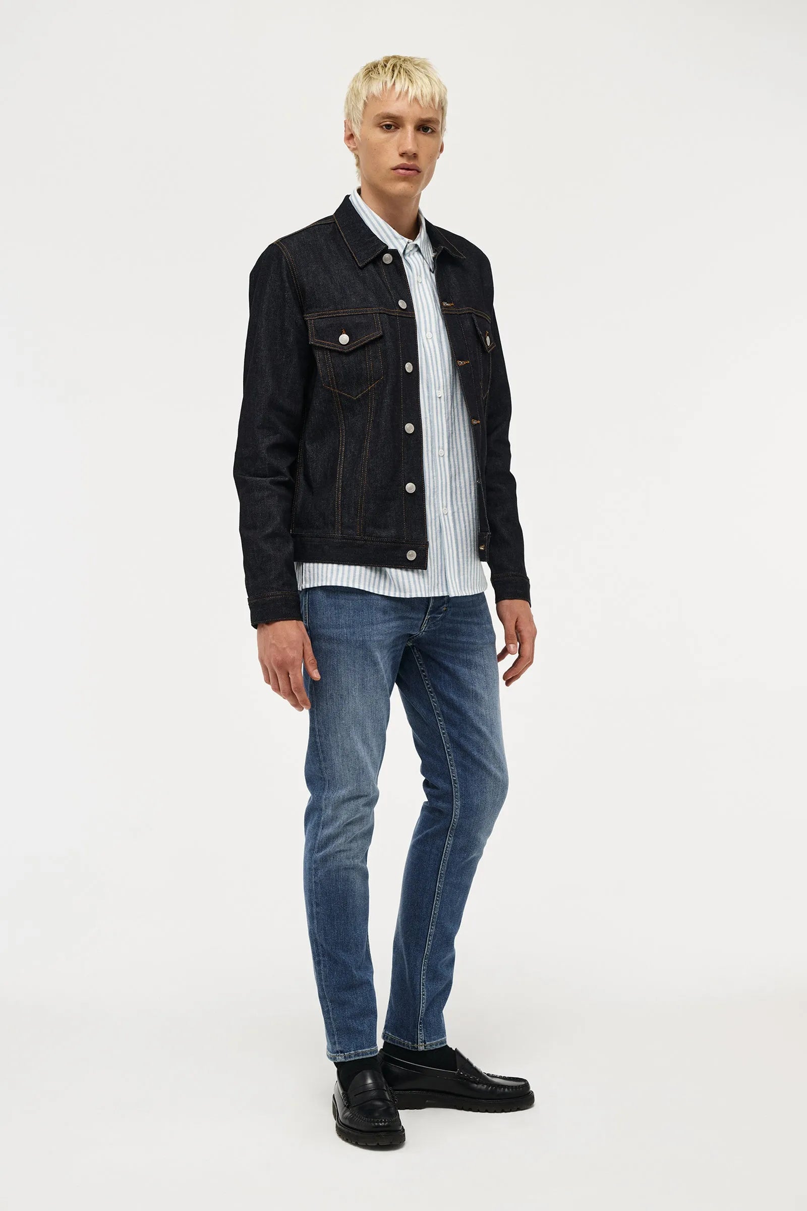 A male model with blonde hair stands against a light gray studio background wearing a black denim jacket over a white and blue striped button-down shirt, blue jeans, and black leather shoes.