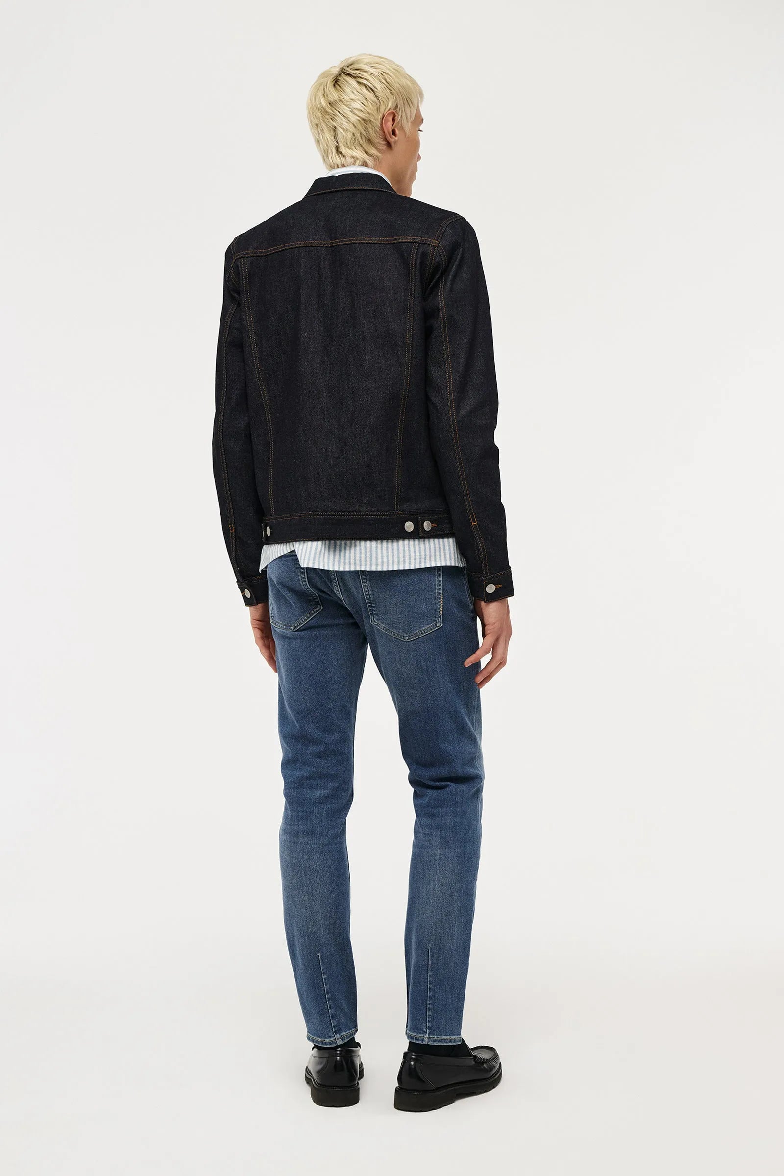 A man viewed from behind wearing a black denim jacket, a white and blue striped shirt, blue jeans, and black shoes, standing against a plain white studio background.