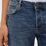 Close-up of a person wearing blue denim jeans with a white top, showing the waistband, front pocket, and button area against a plain light background.