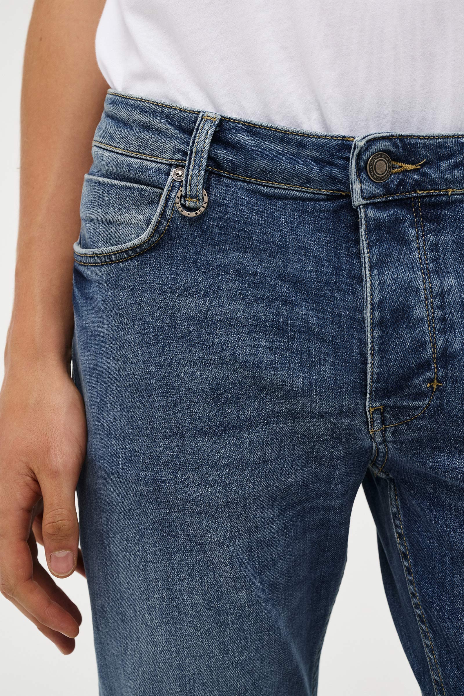 Close-up of a person wearing blue denim jeans with a white top, showing the waistband, front pocket, and button area against a plain light background.